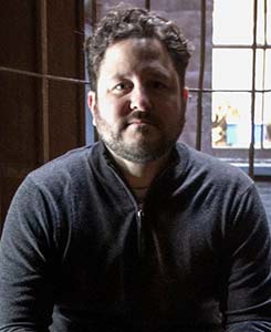 A man with dark curly hair and a beard wearing a dark grey zip-up sweater.