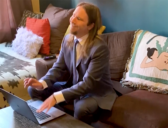 A man with long hair wearing a grey suit sits on a brown couch and types on a laptop.
