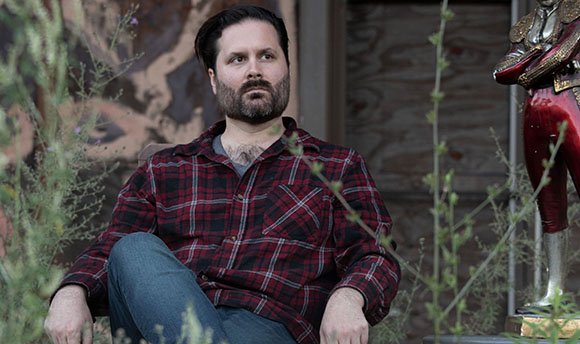 A bearded man in a red plaid shirt sits amidst tall weeds and greenery.