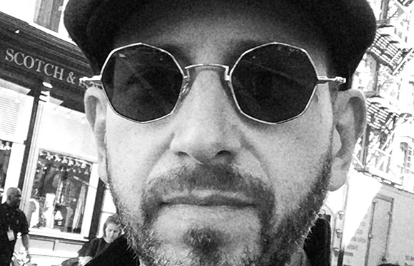Black and white close-up of a man with a beard wearing dark octagonal sunglasses.