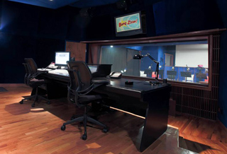 A dark control room with a large mixing desk, computer monitors, and a window looking into a recording booth.