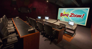 A dark production room with long desks, computer monitors, and a large screen displaying 'Bang Zoom!'.