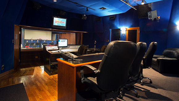A recording studio control room with blue walls, a large mixing console, and black leather office chairs.