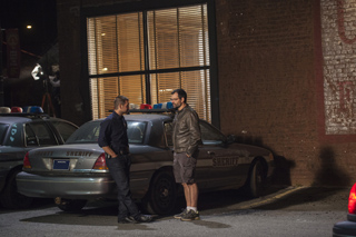 A man in a dark uniform leans against a sheriff's car while talking to a man in a leather jacket.