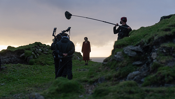 A film crew with a camera and boom mic filming an actress in a red dress on a grassy cliff at sunset.