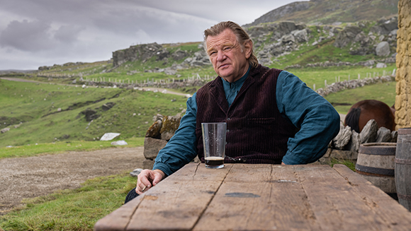A man in a blue shirt and dark velvet vest sits at a wooden table in a hilly, green landscape.