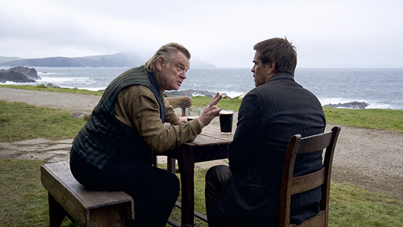 Two men sit at a wooden table outdoors overlooking a cloudy coastline and the ocean.