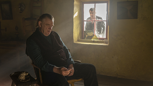 An older man sits in a chair in a dark room as a younger man looks through a window.