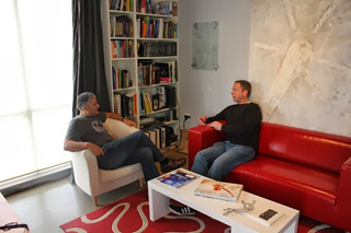 Two men sit on a white armchair and a red sofa in a room with a large bookshelf.