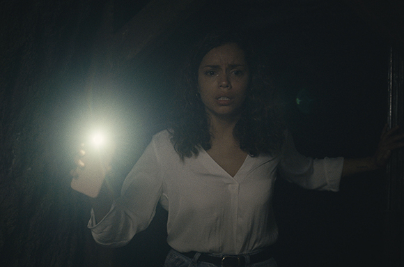 A woman in a white shirt holds a bright flashlight in a dark, shadowy space.