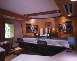 A large mixing console sits in a recording studio with monitors, speakers, and two black office chairs.