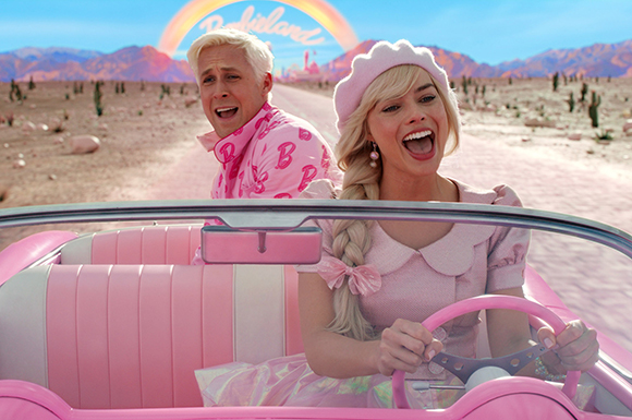 A blonde woman in a pink dress and beret laughs while driving a pink convertible through a desert landscape.