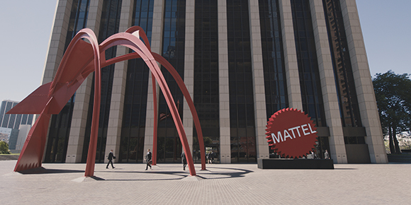 A large red abstract sculpture stands in a plaza before a tall office building with a Mattel sign.
