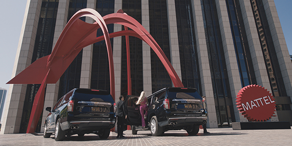 Two black SUVs parked before a large red arch sculpture and a building with a Mattel sign.