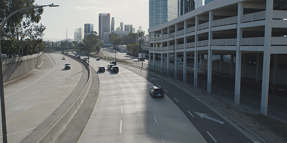 A highway with several cars driving alongside a large concrete parking garage under a city skyline.