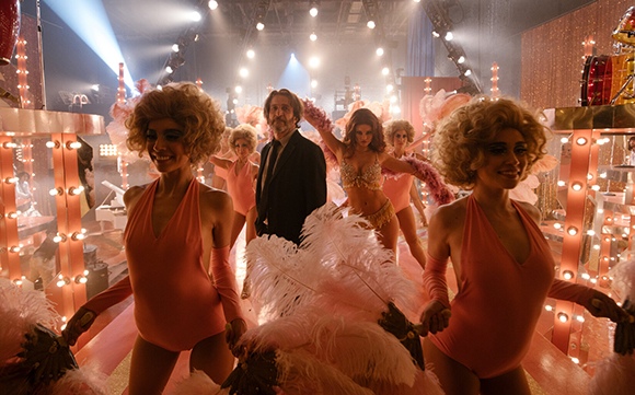 Performers in pink leotards and feather boas pose on a brightly lit stage with a man in a suit.