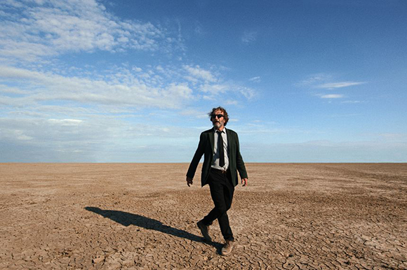 A man in a dark suit and sunglasses walks across a vast, cracked, dry desert under a blue sky.
