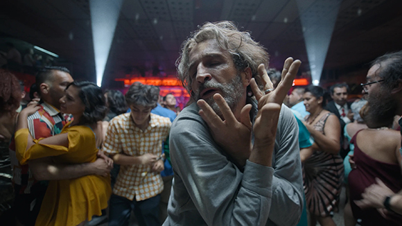 A man with graying hair and a beard holds his hands up in a crowded, dimly lit room.