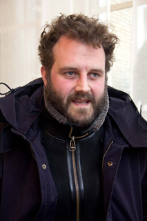 A man with curly hair and a beard wearing a dark zip-up jacket.