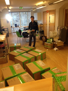 A man stands in a brightly lit room surrounded by several large cardboard boxes taped with green patterned tape.