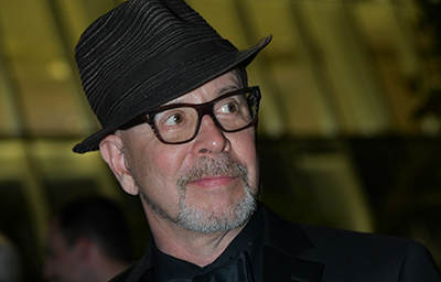 A man with a grey beard, wearing a dark fedora and glasses, looking off-camera.