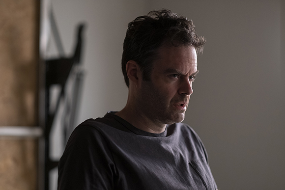 A man with dark hair and a dark t-shirt looks to the side with a concerned, intense expression.