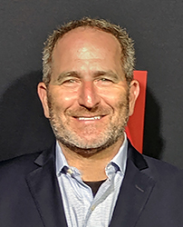 A smiling man with short, curly hair and a beard wearing a blue blazer and light blue shirt.