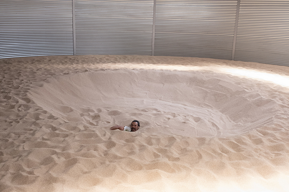 A man lies in a large, shallow pit of sand inside a room with corrugated metal walls.