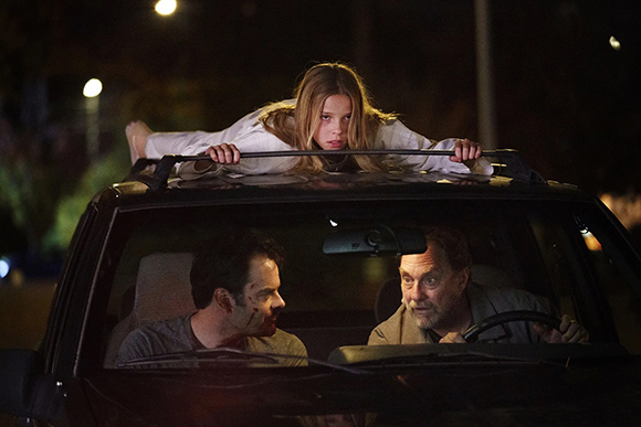 A young girl leans over a car sunroof at night while two men sit in the front seats.