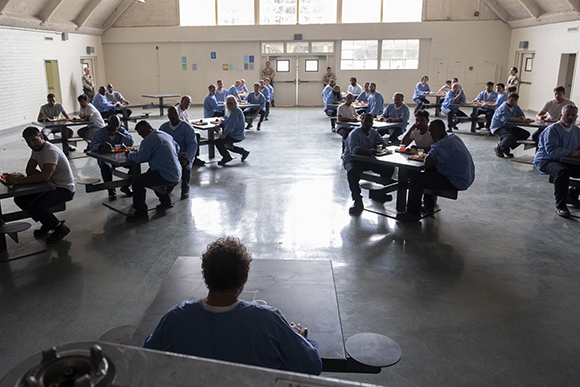 People in blue scrubs sit at dark tables in a large, bright communal dining room with concrete floors.