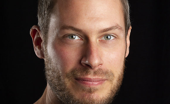 Close-up portrait of a man with light eyes and a short beard against a black background.