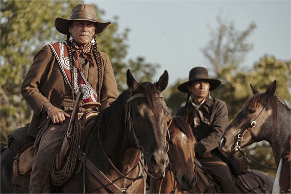 Two people in western attire riding horses through a field with trees in the background.