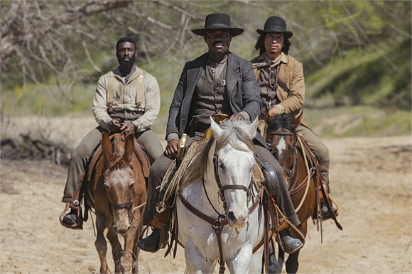 Three men in period clothing riding horses through a dusty, outdoor landscape.