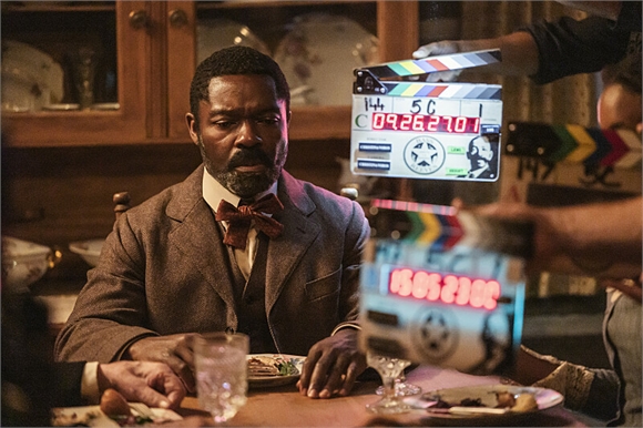 A man in a brown suit and bowtie sits at a table behind a film clapperboard on a movie set.