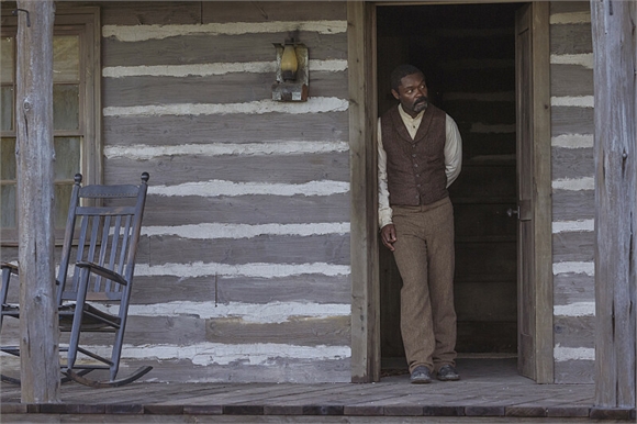A man in a brown vest and trousers stands in the doorway of a rustic log cabin next to a rocking chair.