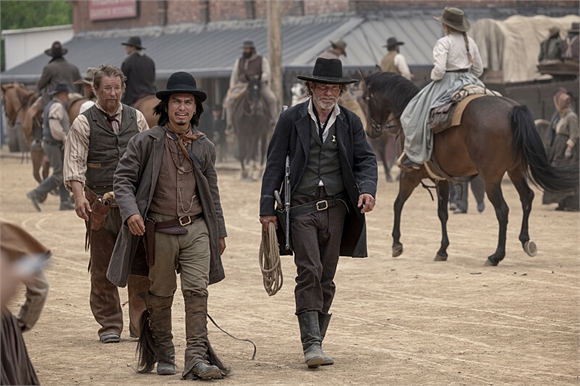 Men in 19th-century western attire walk along a dirt street with riders on horseback in the background.