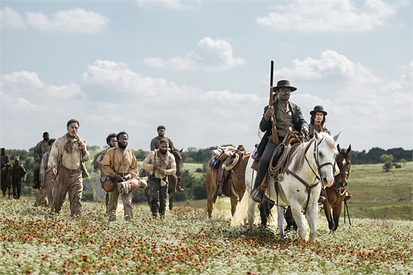 Men in period clothing walk and ride horses through a large field of white and red wildflowers.