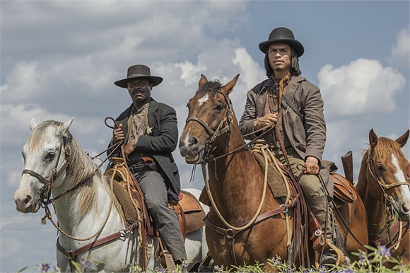 Two men in western attire riding brown and white horses across a field under a cloudy sky.