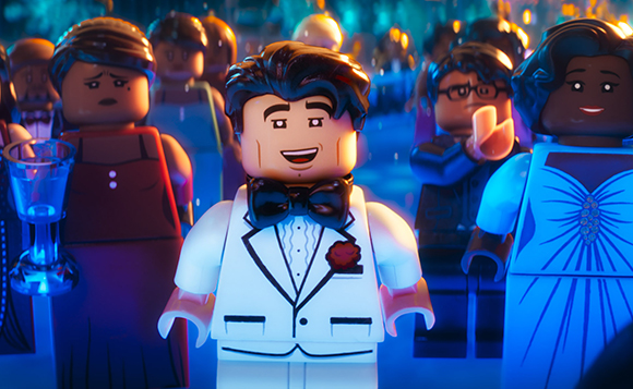A LEGO man in a white tuxedo and black bowtie smiles in a crowd of LEGO figures.