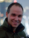 A man with short dark hair smiling while wearing a dark green jacket against a blurred city background.