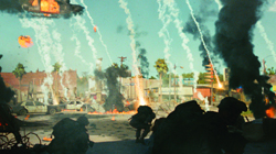 Soldiers crouch in the foreground of a city street under heavy fire and smoke.