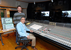 A man sits at a large audio mixing console while another man stands behind him in a recording studio.