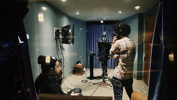 A person in a plaid shirt stands in a recording booth near a microphone and equipment.