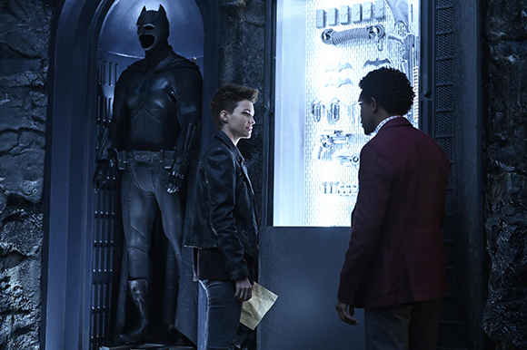 Two young men stand in a dark room looking at a Batman suit inside a glass display case.