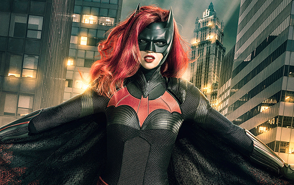 A woman with red hair wearing a black Batwoman suit and mask against a city skyline.