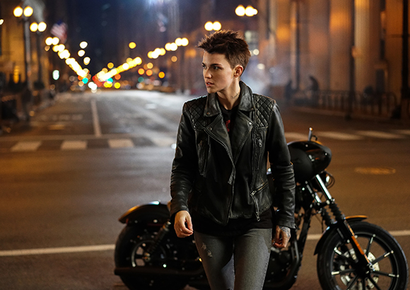 A person with short hair wearing a black leather jacket stands by a motorcycle on a city street at night.