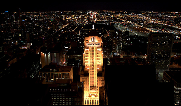 Aerial night view of a city skyline with a central illuminated skyscraper labeled Wayne Enterprises.