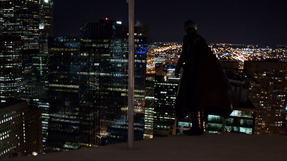 A person in a long dark cloak stands on a rooftop overlooking a brightly lit city at night.