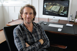 A man with curly hair sits at a wooden desk with his arms crossed in front of a computer monitor.