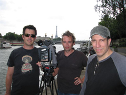 Three men pose outdoors near a river with a professional video camera and the Eiffel Tower in the distance.
